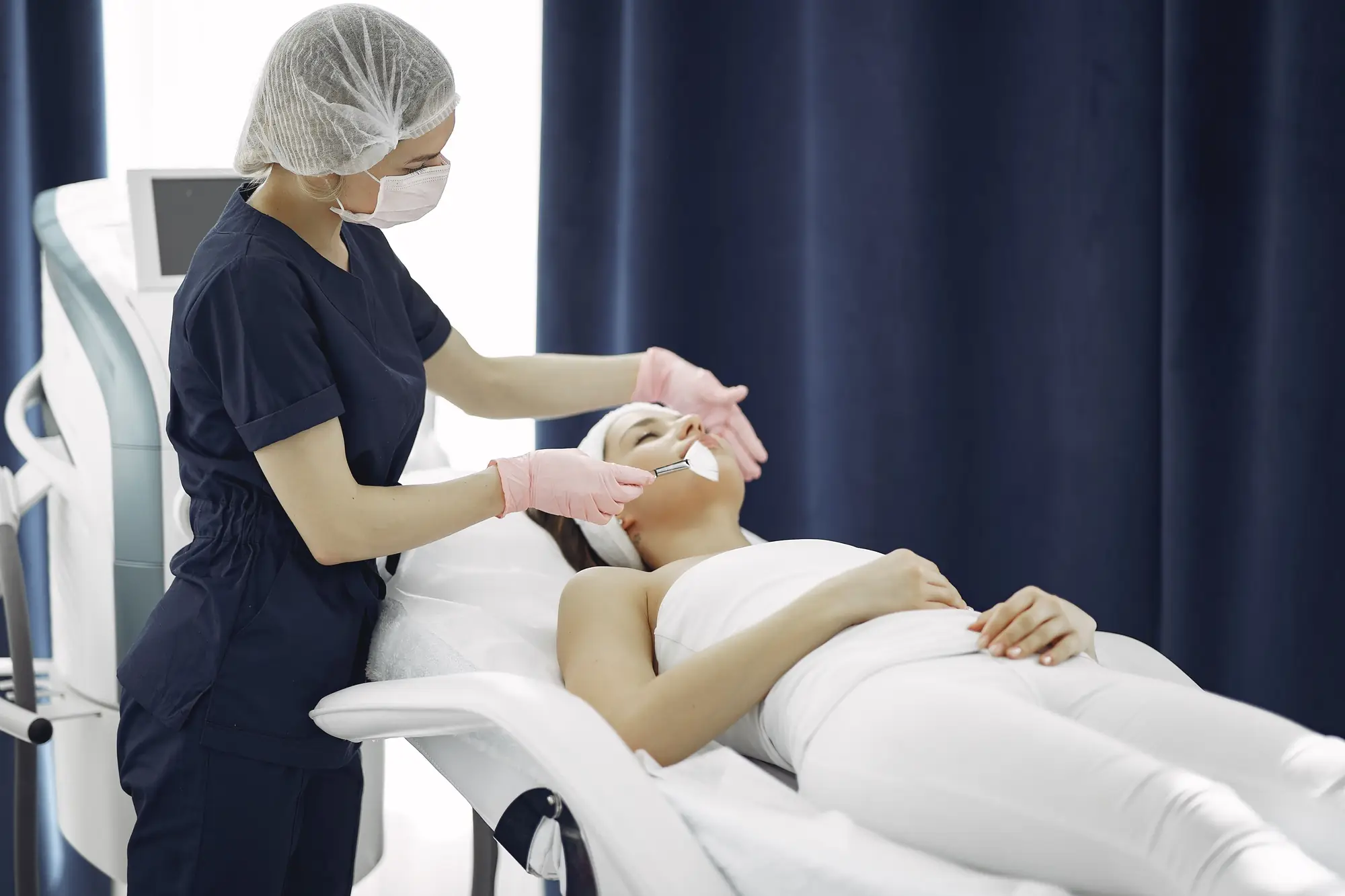 A practitioner treats a patient's skin in a treatment room with a navy curtain.